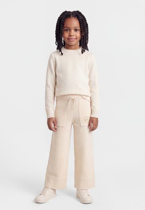 Pige med flettet hår iført en cremefarvet sweater, bukser med vide ben og forlommer samt matchende sneakers, stående mod en ensfarvet baggrund.