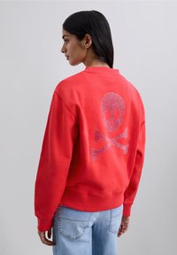 Femme aux longs cheveux noirs portant un sweat-shirt rouge avec un design de skull et os croisés pailleté dans le dos et un jean bleu clair.