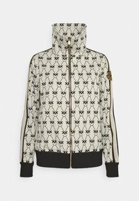 Chaqueta con cremallera en blanco y negro con cuello alto, que presenta un patrón repetido de mariposas, herrajes de cremallera dorados y puños y dobladillo acanalados en negro.