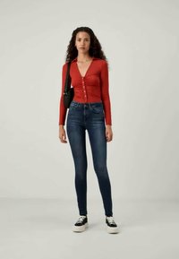 Κόκκινη μακριά μπλούζα με κουμπιά συνδυασμένη με σκούρα μπλε στενά τζιν. Το μοντέλο φοράει μαύρα αθλητικά παπούτσια και κρατάει μία μικρή μαύρη τσάντα ώμου.