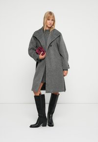 Grauer Wollmantel mit übergroßer Kragen, Ein-Knopf-Verschluss, kombiniert mit einem grauen Kleid und schwarzen knielangen Stiefeln. Burgunderfarbene Clutch in der Hand.