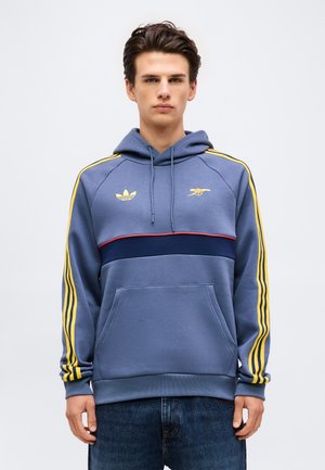 Junger Mann, der einen blauen Hoodie mit gelben Streifen an den Ärmeln, Logos auf der Brust und einem marineblauen horizontalen Streifen vorne trägt, steht vor einem schlichten Hintergrund.