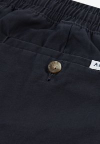 Σκούρο μπλε σορτς με ελαστική μέση, διαθέτοντας ένα καφέ κουμπί και μαλακή βαμβακερή υφή με διακριτική λάμψη.