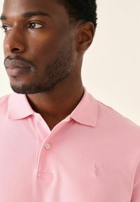 Camisa polo rosa con tejido texturizado, un cuello clásico y un sutil logo bordado en el lado izquierdo del pecho. Cuenta con tapeta de dos botones.
