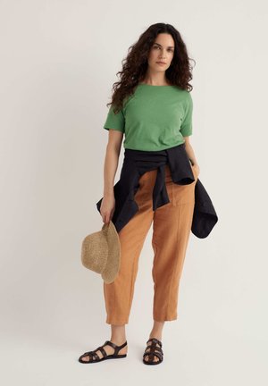 Kvinde med langt krøllet hår iført grøn skjorte, beige bukser, sort jakke bundet om livet, holder en stråhat og har sorte sandaler på.