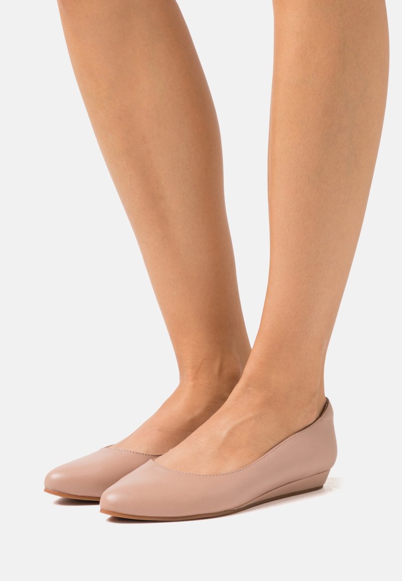 Jambes portant des chaussures compensées fermées couleur nude avec des talons bas sur un fond clair uni.