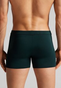 Tmavě zelené přiléhavé boxerky vyrobené z hladkého materiálu, s jednoduchým páskem a bez viditelných vzorů nebo akcentů.