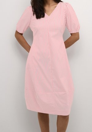 Femme portant une robe rayée verticale rose et blanche jusqu'aux genoux avec des manches courtes bouffantes et un col en V, debout les mains dans le dos.