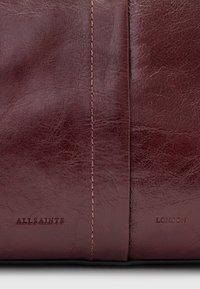 Borsa in pelle bordeaux con una texture liscia, caratterizzata da dettagli cuciti e marchio impressionato "ALLSAINTS LONDON" sulla parte anteriore.