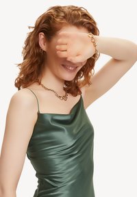 Robe en satin vert avec des bretelles spaghetti et un design drapé. Collier en chaîne dorée avec des maillons entrelacés et boucles d'oreilles en or visibles.