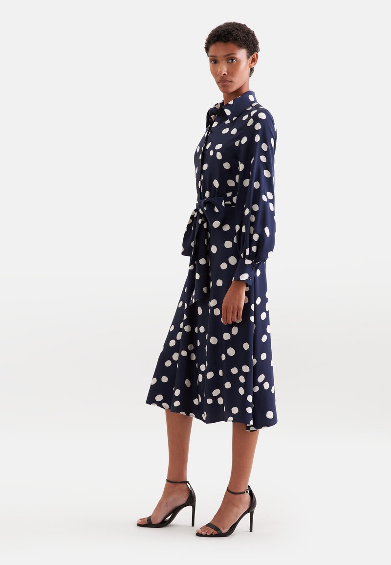 Une femme se tient de profil vêtue d'une robe midi bleu marine à pois blancs, manches longues, col, ceinture à la taille et sandales noires à talons hauts.