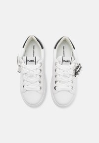 Adidași albi cu un brâu din piele netedă, vârf rotund, șase găuri pentru șireturi și un accent pe călcâi din piele lacuită neagră. Au un logo pe limbă și un fermoar decorativ.