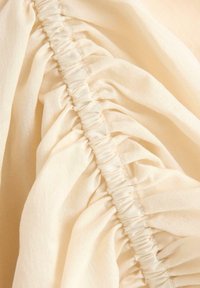 Krämfärgad tyg med insamlad textur, som visar en serie tätt sydda veck som löper vertikalt längs sömmen. Mjukt, slätt material.