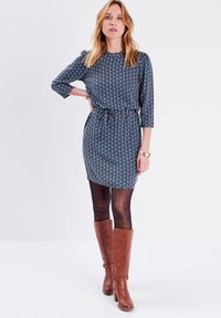 Vestido corto de estampado floral en azul, con mangas tres cuartos y cintura ceñida por un cinturón. Lleva botas marrones hasta la rodilla y medias oscuras.