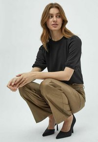 Zwarte korte mouw top, bruine wijde broek, en zwarte hoge hakken. Model in hurkhouding met handen op de knieën, met een neutrale achtergrond.