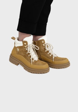 Personne portant des bottines lacées beige avec doublure en polaire blanche et semelles épaisses en caoutchouc, associées à un pantalon noir à revers.