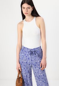 Fehér ujjatlan bodysuit kék mintás nadrággal, amelynek derékrészén húzózsinór található. A modell egy barna táskát tart.