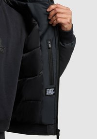 Mustapufferi takki, jossa on sileä pinta ja tikattu vuori, varustettuna vetoketjullisella taskulla ja sisällä logolla varustetulla tarralla.