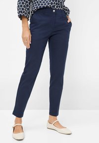 Marineblaue maßgeschneiderte Hose aus Baumwolle, mit schlankem Schnitt, flachem Vorderteil und Knopfverschluss. Kombiniert mit cremefarbenen flachen Schuhen.