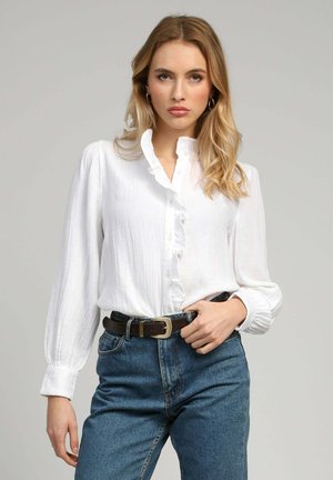 Femme blonde portant un chemisier blanc à volants rentré dans un jean bleu taille haute avec une ceinture noire, debout devant un fond gris uni.