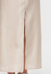 Μπεζ μίντι φούστα με ανοίγμα στο μπροστινό μέρος που αποκαλύπτει τα κάτω πόδια και ασημένιο βραχιολάκι στον αστράγαλο, σε λευκό φόντο.