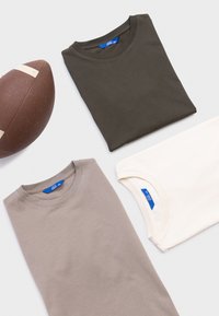 Drei gefaltete Baumwoll-T-Shirts in Olivgrün, Beige und Cremeweiß. Runde Ausschnitte mit genähten Nähten und sichtbaren blauen Markenetiketten. Ein brauner Fußball in der Nähe.