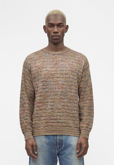 Jeune homme aux cheveux blonds courts portant un pull texturé multicolore et un jean bleu clair sur un fond blanc uni.