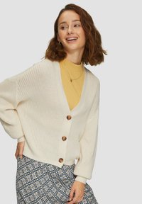 Cremefarbener offener Strickcardigan mit Holzknöpfen, langen Ärmeln und lockerem Schnitt, getragen über einem gelben Rippenoberteil und einem Rock mit floralem Muster.