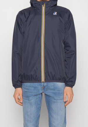 Person trägt eine marineblaue Kapuzenjacke mit gelb-weiß gestreiftem Reißverschluss und einem K-Way-Logo auf der linken Brust, kombiniert mit blauen Jeans.