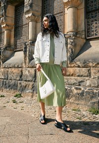 Giacca di jeans bianca sopra un pullover verde, gonna verde plissettata, sandali neri con cinturini incrociati e una borsa bianca con manico superiore.