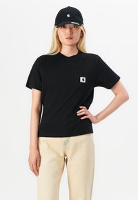 Black cotton t-shirt with short sleeves and a pocket featuring a logo. Paired with beige pants and a matching black cap.