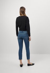 Sudadera negra, jeans ajustados de color azul con un largo ligeramente recortado y zapatos negros tipo slip-on con detalles dorados. Vista trasera, fondo neutro.