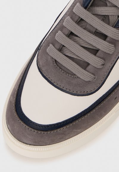 Adidași gri din suede cu un panou crem, linii de accent bleumarin și șireturi plata gri, având cusături texturate pe margini.