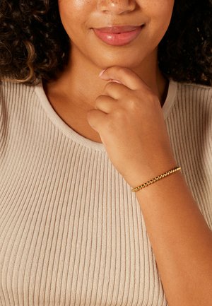 Gold beaded bracelet on wrist; smooth, round beads create a classic design. Worn with a light beige ribbed top.