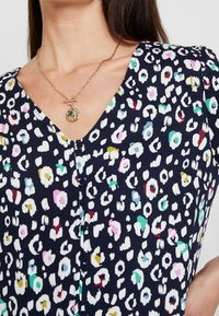 Blouse navy avec un col en V, ornée d'un imprimé animal coloré en blanc, rose, vert et jaune, et de petits boutons sur le devant.