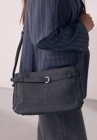 Melnas krokodila raksta soma no teksturētas ādas, ar platu siksnu, sprādzes detaļu un taisnstūrveida formu.