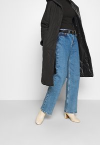Abrigo negro acolchado largo, top negro ajustado, vaqueros de pierna ancha azul claro, cinturón negro con hebilla dorada y botines de tacón beige.