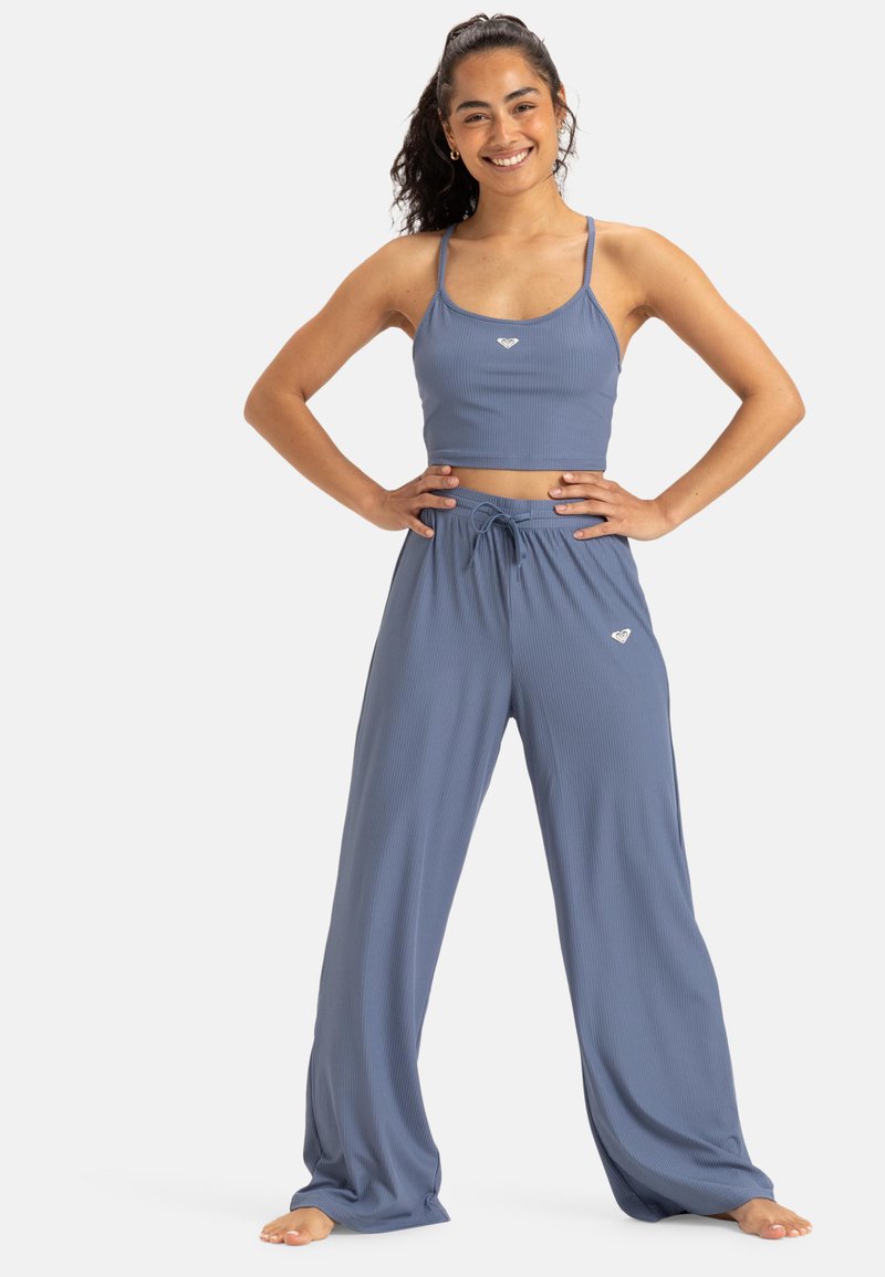 Femme souriante en top court côtelé bleu et pantalon large assorti, pieds nus, les mains sur les hanches, contre un fond blanc.