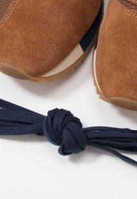 Baskets en daim brun avec une semelle extérieure en caoutchouc et des lacets bleu marine, présentant un détail de lacet noué serré à l'avant.