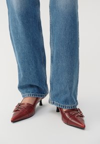Zapatos de punta fina de cuero rojo con un pequeño tacón, que presentan costuras horizontales y dos hebillas plateadas, usados con jeans de mezclilla azul.