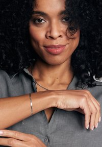 Srebrna metaliczna bransoletka na nadgarstku, noszona z szarym t-shirtem; kręcone włosy otaczające twarz, delikatny makijaż podkreślający rysy.