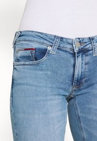 Detailní záběr světle modrých džínů s předními kapsami, poutky na opasek a kovovým knoflíkem, nošených s bílým ribbovaným topem.