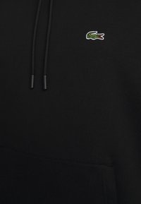 Sudadera negra con bolsillo tipo canguro, cordones y un pequeño logotipo de cocodrilo verde bordado. Textura de tela suave.