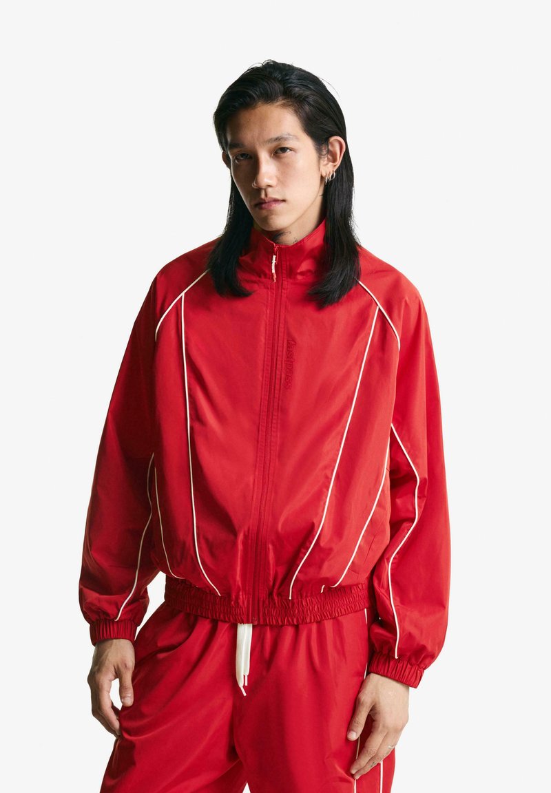 Red windbreaker jacket featuring white piping, high collar, elastic cuffs, and waistband. Paired with matching pants for a co-ordinated look.