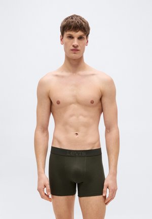 Άνδρας χωρίς μπλούζα στέκεται αντικρίζοντας μπροστά, φορώντας σκουροπράσινα μποξέρ Levi's, μπροστά σε απλό ανοιχτό φόντο.