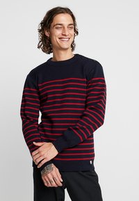 Granatowy sweter w czerwone poziome paski, pleciona tekstura, okrągły dekolt oraz guziki na ramieniu. Dopasowany krój z długimi rękawami.