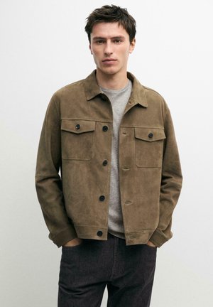 Giovane uomo con giacca marrone in camoscio sopra una maglietta grigia, con le mani nelle tasche, in piedi davanti a uno sfondo chiaro uniforme.