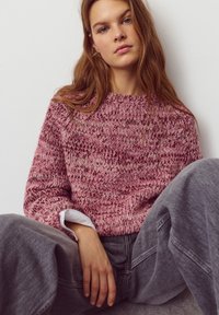 Pulover tricotat în tonuri de roz și burgund, cu gât rotund și model texturat. Asortat cu pantaloni gri, cu croială lejeră, purtat peste o cămașă albă.