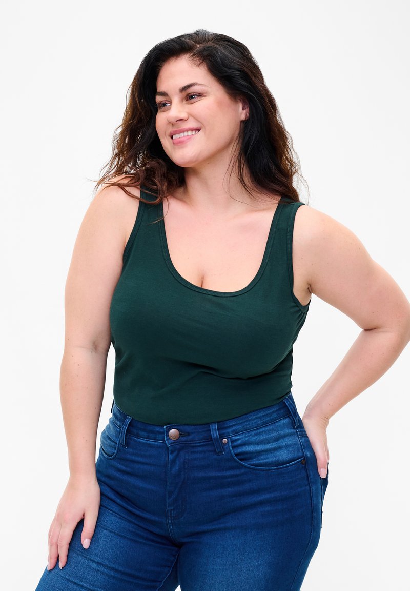 Tume roheline tank top, mis on valmistatud pehmest materjalist ja millel on ümar kaelus. Kombineeritud kõrge vöökohaga siniste teksadega. Sile tekstuur, keha kohandav disain.