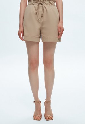 Mujer que lleva shorts paperbag de talle alto beige con cordón y tacones nude con tiras en el tobillo, de pie frente a un fondo liso.